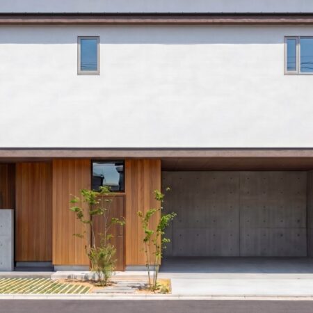 Front view of a modern house with a white stucco wall brown framing and a wooden entry door concrete base and greenery nearby 神戸市の注文住宅