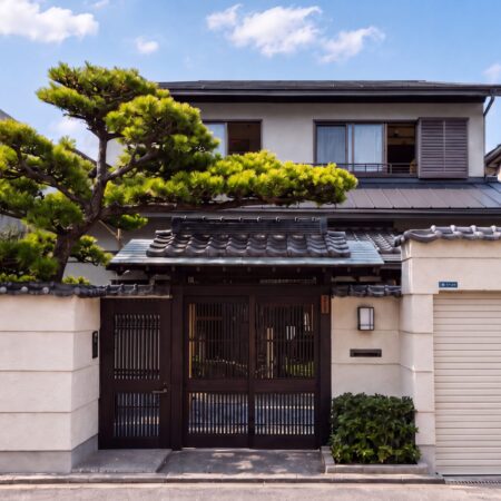 Japanese style house with a dark wooden gate tiled roof and manicured pine tree in the front yard against a blue sky and neighboring modern buildings 神戸市の注文住宅